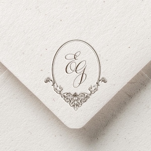 Puede incluir: Primer plano de una esquina de sobre color crema con un monograma plateado dentro de un marco ovalado. El monograma incluye las letras "E" y "G" en una escritura elegante, con adornos decorativos. El texto "KATAKALA STUDIO" está en la parte inferior.