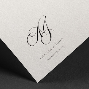 Può includere: Un invito di nozze con un monogramma calligrafico delle lettere "A" e "J" in inchiostro nero. Il testo "Amanda & John" e "September 25, 2025" è stampato sotto il monogramma.