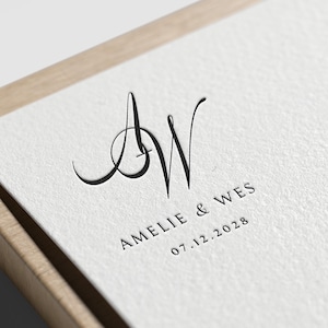 May include: Close-up of a wedding invitation with the initials "AW" in elegant black script. The names "Amelie & Wes" and the date "07.12.2028" are printed below. The invitation is set in a wooden frame.