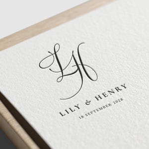 May include: A close-up of a white card with a black, elegant monogram of the letters L and H. The names "LILY & HENRY" and the date "18 SEPTEMBER 2028" are printed below the monogram in a serif font.