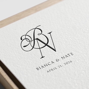 Op de afbeelding: Een witte kaart met een zwart monogram van de letters "B" en "N" in een schrijfletter. De tekst "Bianca & Nate" en "April 21, 2026" staat onder het monogram.