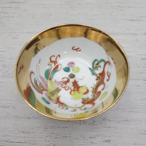 Peut inclure: Un petit bol en porcelaine blanche avec un bord doré et un motif de dragon coloré. Le bol est décoré de peinture rouge, verte, jaune et bleue.