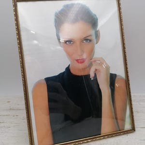 May include: A rectangular, gold-toned picture frame with a portrait of a woman. The frame has a detailed, textured border. The woman in the portrait has fair skin, red lipstick, and is wearing a black top.