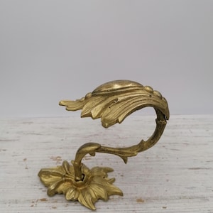 May include: A gold-colored, ornate curtain tieback with a floral design. The tieback features a stylized flower base, a curved stem, and a leaf-shaped holder. The metal has a slightly aged appearance.
