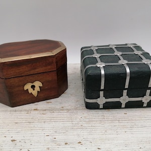 May include: Two decorative boxes. One is a dark wood, octagonal shape with brass trim and a leaf-shaped clasp. The other is a square, dark green box with silver metal gridwork and floral accents. Both are closed.