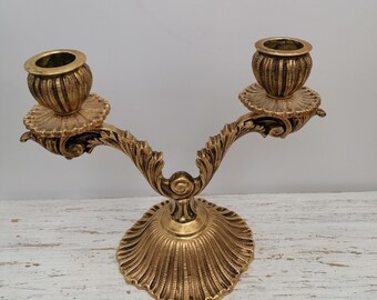Puede incluir: Un candelero de metal dorado con dos portavelas. El candelero tiene un diseño decorativo y ornamentado con una base festoneada.