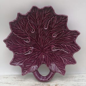Peut inclure: Un plat de service en céramique violet foncé en forme de grande feuille d'érable texturée. La pièce présente une finition émaillée brillante, des détails de nervures en relief sur la surface et une poignée circulaire intégrée à la tige.