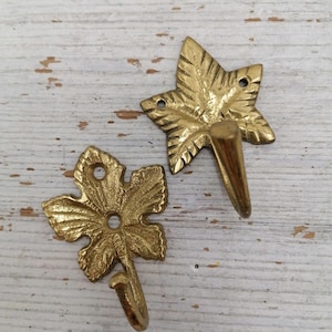 Op de afbeelding: Twee messing wandhaken in de vorm van een blad. De haken zijn gedetailleerd met bladnerven en hebben een warme, gouden kleur. Ze zijn ontworpen om aan een muur te worden bevestigd om items op te hangen. De haken zijn van metaal.