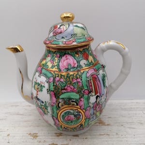 May include: A white porcelain teapot with a lid, spout, and handle, decorated with vibrant floral and figural patterns. The teapot has gold detailing on the spout, lid knob, and handle. The design incorporates pink, green, and gold colours.