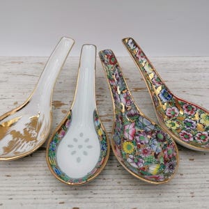 Könnte beinhalten: Vier Suppenlöffel aus Porzellan mit unterschiedlichen Designs. Zwei Löffel zeigen komplizierte, bunte Blumenmuster mit Goldrand. Ein Löffel hat ein weißes Inneres mit einem zentralen Blumenmotiv und Goldkanten. Ein Löffel zeigt eine Goldlandschaft.