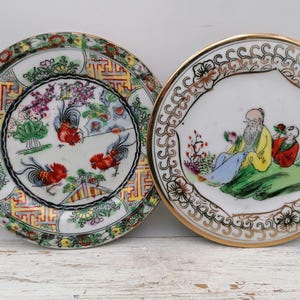 Peut inclure: Deux assiettes décoratives en porcelaine. L'une présente un motif de coq avec des accents rouges, blancs et verts. L'autre assiette a un bord doré et représente une figure assise avec deux autres. Les deux assiettes ont des motifs complexes.