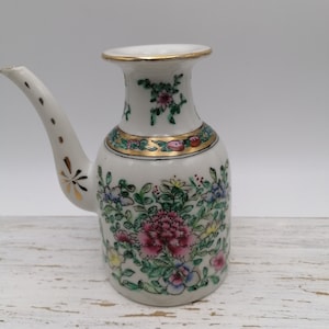 Può includere: Una teiera in ceramica bianca con un design floreale in tonalità rosa, verde e giallo. La teiera ha un beccuccio e un manico. La teiera è decorata con accenti dorati.
