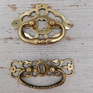 Può includere: Due maniglie antiche per cassetti in ottone. La maniglia superiore ha un design traforato decorativo con una maniglia curva e tre pomelli decorativi. La maniglia inferiore ha un buco della serratura e una maniglia ritorta.