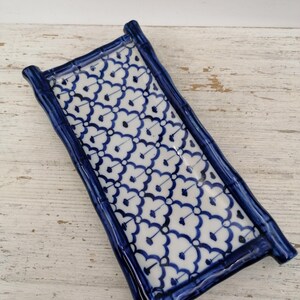 May include: A rectangular ceramic tray with a blue and white floral pattern. The tray has a bamboo-style frame in a deep blue colour. The white surface is decorated with a repeating blue floral design. The tray is placed on a distressed white wooden surface.