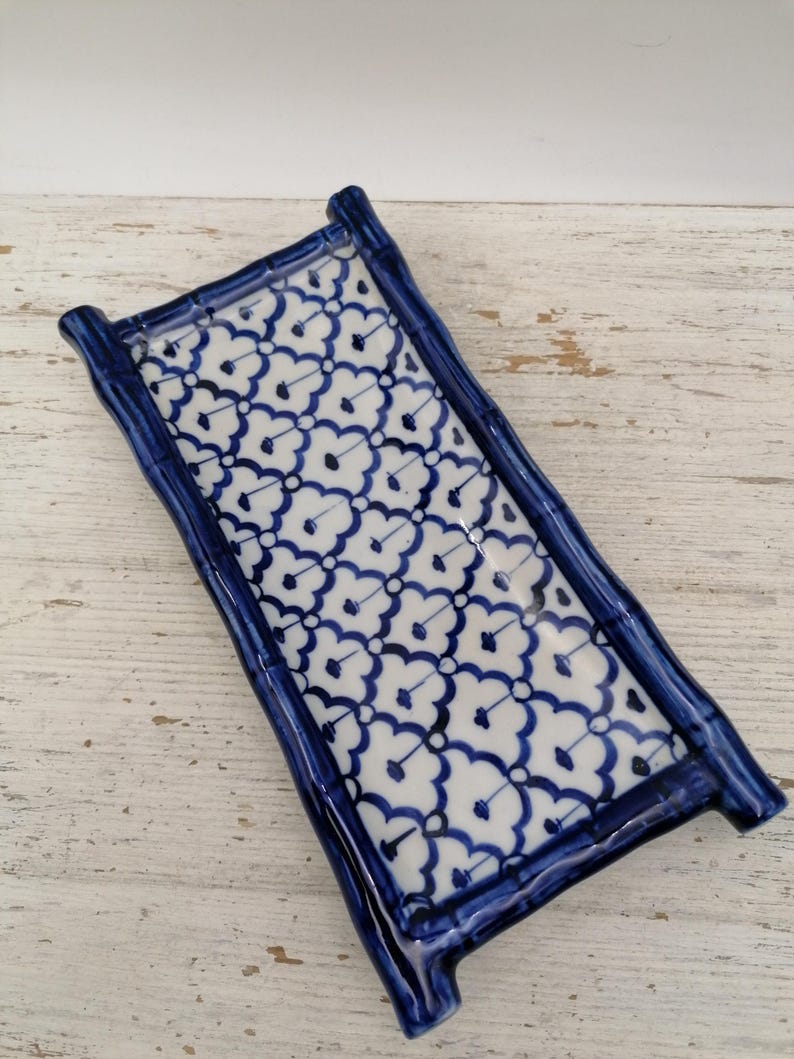 May include: A rectangular ceramic tray with a blue and white floral pattern. The tray has a bamboo-style frame in a deep blue colour. The white surface is decorated with a repeating blue floral design. The tray is placed on a distressed white wooden surface.