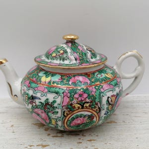 May include: A porcelain teapot featuring a white base with intricate, colourful floral and butterfly patterns in shades of pink, green, and gold. The teapot includes a matching lid with a gold-toned knob, a curved spout, and a handle with gold trim.