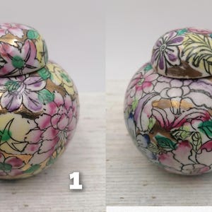 May include: Two small, round porcelain jars with lids. Both jars are decorated with a floral pattern in shades of pink, yellow, green, and gold. The jars are sitting on a white surface.