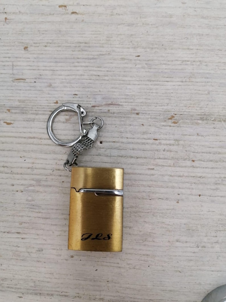 Puede incluir: Un encendedor de llavero de color dorado con una cadena y un anillo plateados. El encendedor tiene la inscripci&oacute;n "J.L.S." en la parte delantera.