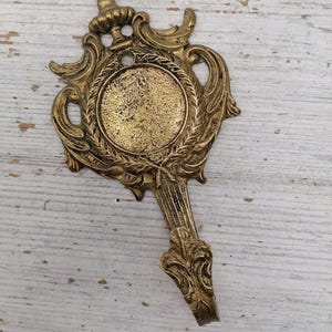 May include: Ornate, antique brass-colored wall hook with a decorative, symmetrical design. The hook features a central circular frame, surrounded by intricate scrollwork and a crown-like top. The hook is likely for decorative purposes.