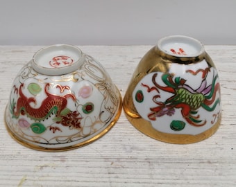 May include: Two small white porcelain bowls with gold trim and painted red, green, and gold dragons. The bowls are on a white wooden surface.