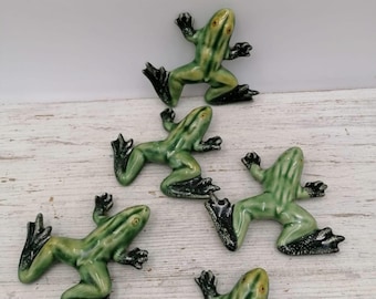Puede incluir: Figuras de rana de cerámica verde con patas negras y ojos amarillos, dispuestas sobre una superficie de imitación madera clara. El texto "The price is for 1 frog" está en un rectángulo blanco.