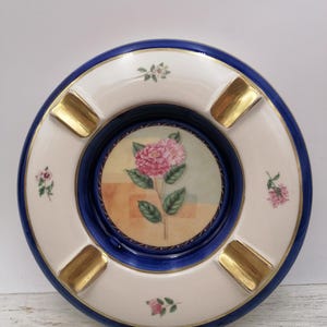 Op de afbeelding: Ronde keramische asbak met een blauwe rand en een wit midden. Het midden heeft een bloemenontwerp met een roze bloem. Goudkleurige rustplaatsen zijn rond de rand geplaatst. Kleine roze bloemaccenten zijn ook aanwezig.