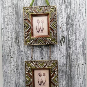 Peut inclure: Deux cadres photo carrés décoratifs avec une impression botanique de tulipes. Les cadres présentent un motif géométrique marron et vert avec des accents de cristaux transparents. Suspendus par un ruban et une chaîne verts.