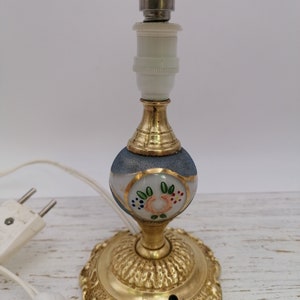 May include: A small table lamp with a green lampshade and a white base with painted floral details. The base is made of brass and has a decorative design. The lamp is wired with a plug.