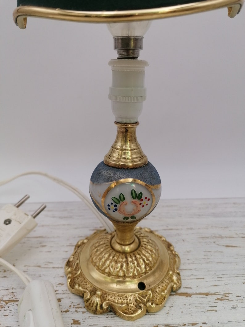 May include: A small table lamp with a green lampshade and a white base with painted floral details. The base is made of brass and has a decorative design. The lamp is wired with a plug.