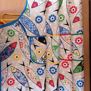 Peut inclure: Un tablier de cuisine arborant un motif répétitif de poissons stylisés aux couleurs variées, notamment rouge, bleu, vert et jaune, entremêlés de motifs floraux sur un fond beige clair. Le tablier présente un liseré bleu et des liens jaunes.