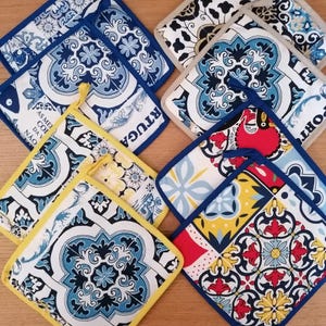 May include: A collection of square fabric pot holders featuring various tile-inspired patterns in blue, white, yellow, and red. Some items have blue or yellow fabric trim and hanging loops, while others feature beige trim and zippers.