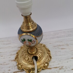 May include: A gold and white table lamp with a floral design. The lamp has a white cord and a white ceramic base with a gold trim. The lamp is on a white wooden surface.