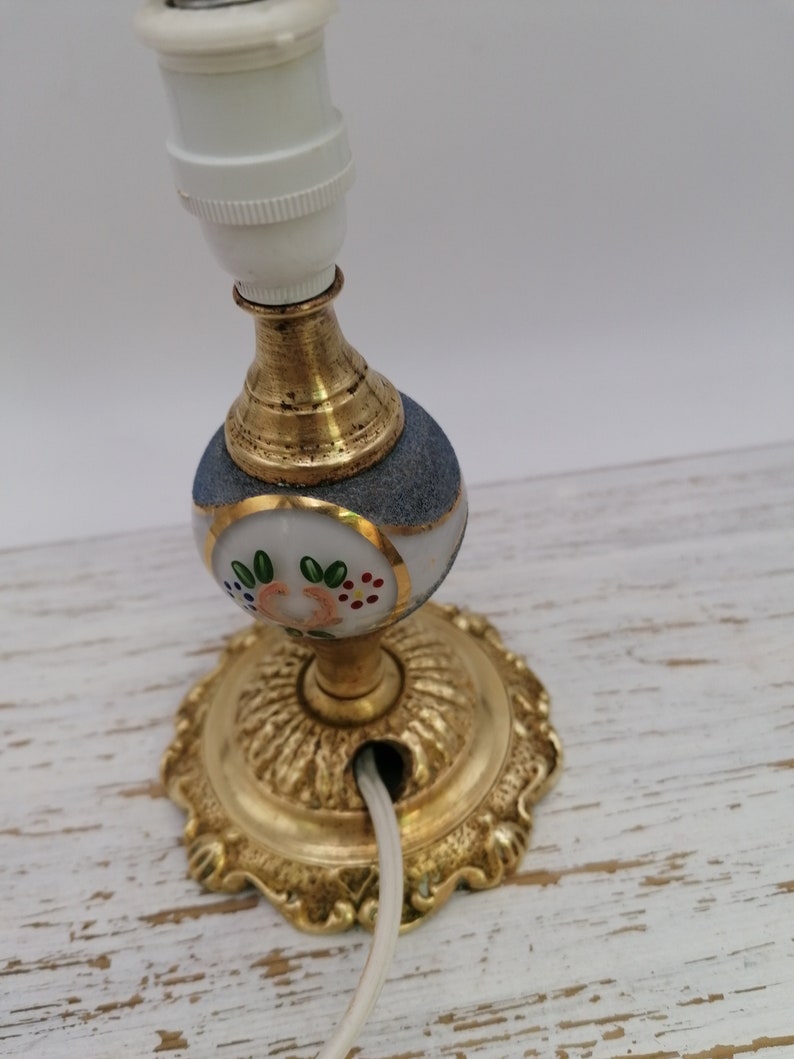 May include: A gold and white table lamp with a floral design. The lamp has a white cord and a white ceramic base with a gold trim. The lamp is on a white wooden surface.
