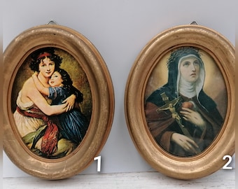 May include: Two oval-shaped framed prints. One depicts a woman embracing a child, the other a religious figure holding a crucifix. Both prints are in gold-colored frames and are mounted on a white surface.