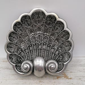 May include: A silver-toned metal dish shaped like a scallop shell. The surface features intricate embossed scrollwork, floral patterns, and beaded lines radiating from a central smooth, rounded base flanked by two prominent spiral curls.