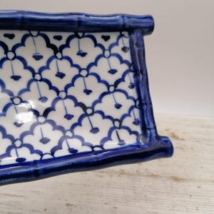 May include: A rectangular ceramic dish with a blue and white floral pattern. The dish has a raised, curved edge and is painted with a repeating design of blue flowers on a white background. The edges are a solid, dark blue.