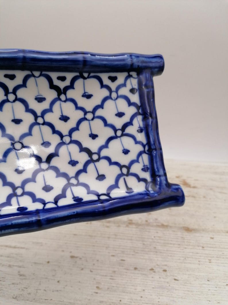 May include: A rectangular ceramic dish with a blue and white floral pattern. The dish has a raised, curved edge and is painted with a repeating design of blue flowers on a white background. The edges are a solid, dark blue.