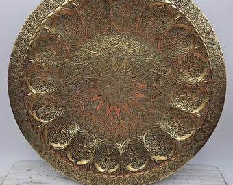 May include: A large, round, antique brass tray with intricate embossed floral and geometric patterns. The tray features a central starburst design and a decorative border. The color is a warm gold with darker accents, suggesting age and use.