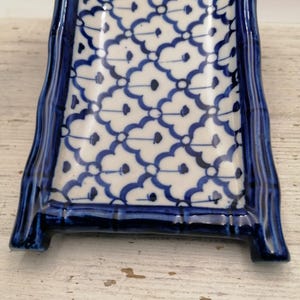 May include: A rectangular ceramic serving dish with a blue and white floral pattern. The dish has a raised, curved border and is supported by two small feet. The design features a repeating pattern of blue floral motifs on a white background.