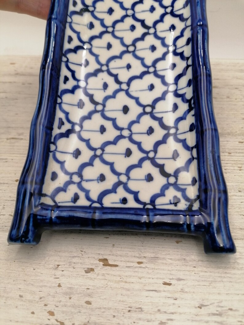 May include: A rectangular ceramic serving dish with a blue and white floral pattern. The dish has a raised, curved border and is supported by two small feet. The design features a repeating pattern of blue floral motifs on a white background.