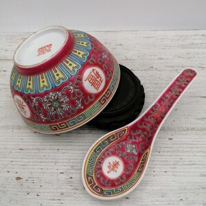 May include: A red and white porcelain bowl and spoon set with detailed floral and geometric patterns. The bowl sits on a black wooden stand. The spoon and bowl feature Chinese characters.