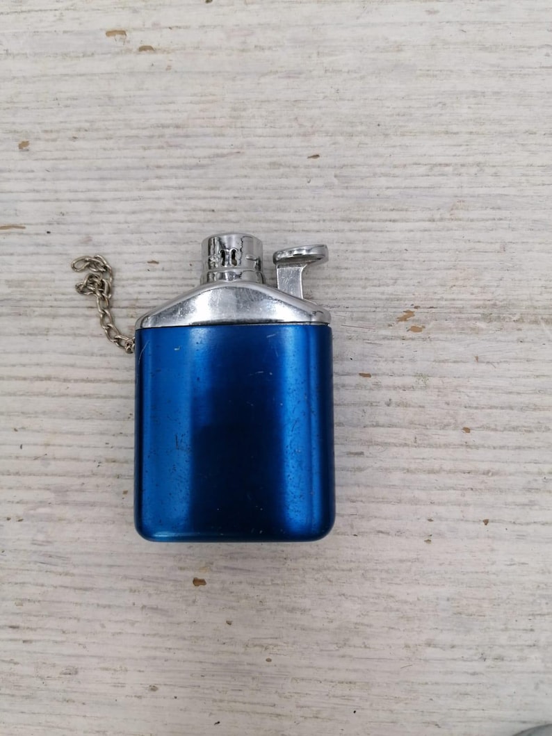 Puede incluir: Un encendedor de metal azul con una parte superior plateada y una cadena adjunta.
