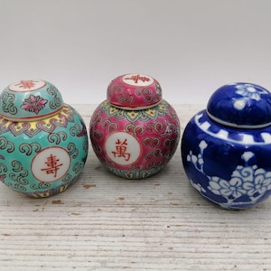 Puede incluir: Tres tarros decorativos de cerámica con tapas. Uno es turquesa con detalles rojos y amarillos y caracteres chinos. El segundo es rosa con detalles amarillos y caracteres chinos. El tercero es azul con diseños florales blancos.