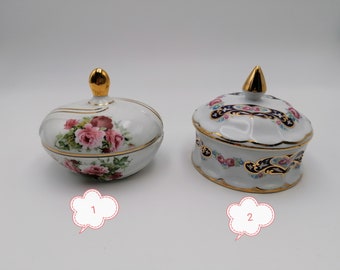May include: Two white porcelain trinket boxes with gold trim and floral designs. The box on the left has a pink rose design. The box on the right has a blue and gold paisley design.