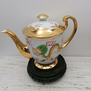 Op de afbeelding: Een witte porseleinen theepot met een gouden rand en een groen en rood drakenontwerp. De theepot staat op een zwarte houten standaard.