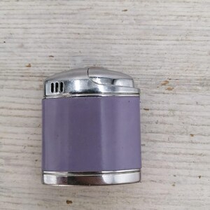 Puede incluir: Un encendedor plateado y morado con forma rectangular redondeada. El encendedor tiene una parte superior plateada y un cuerpo morado.