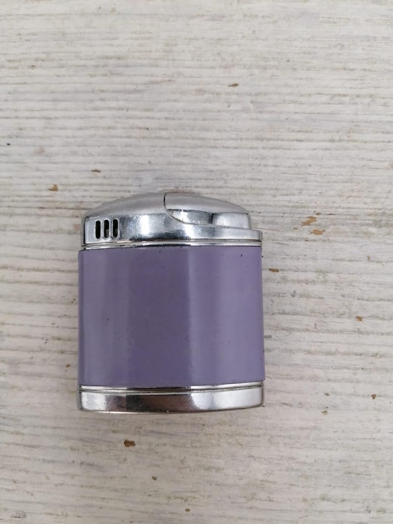 Puede incluir: Un encendedor plateado y morado con forma rectangular redondeada. El encendedor tiene una parte superior plateada y un cuerpo morado.
