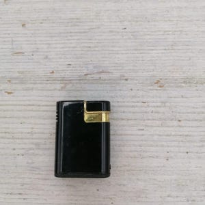 Puede incluir: Un encendedor negro con un borde dorado. El encendedor est&aacute; sobre una superficie de madera blanca.