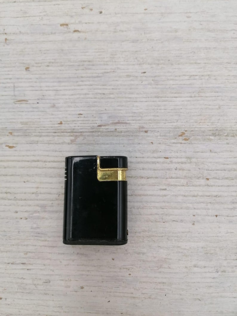 Puede incluir: Un encendedor negro con un borde dorado. El encendedor est&aacute; sobre una superficie de madera blanca.