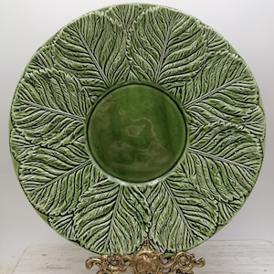 Peut inclure: Une grande assiette en céramique verte avec un motif en relief de feuilles de chou. L'assiette est sur un support en métal doré.